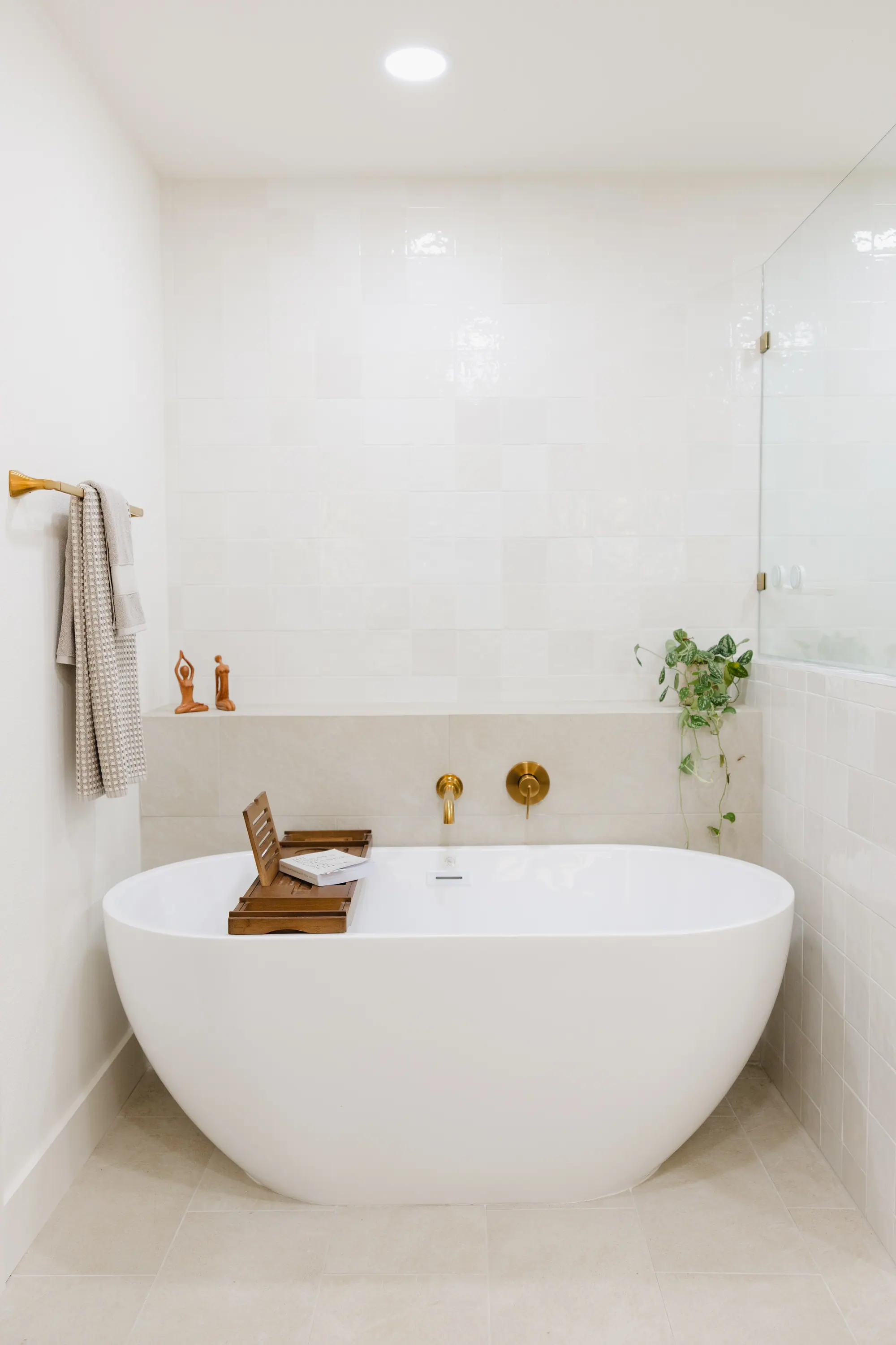 A finished primary bath: a white freestanding tub below a stone ledge, square white wall tile, brushed-gold wall-mounted tub fittings and a glass shower return.