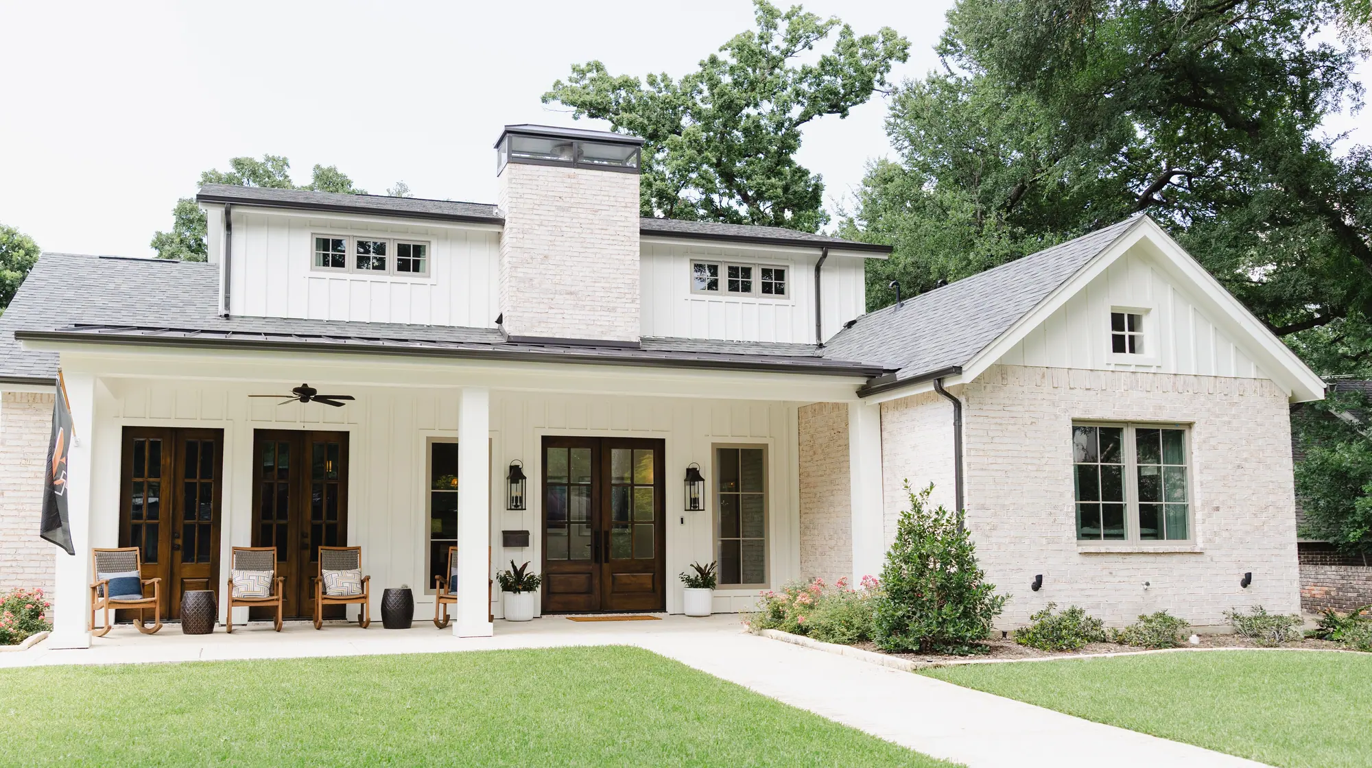 The finished house from the front yard: white board-and-batten, a painted brick gable wing and chimney, a metal porch roof, dark wood double doors and rocking chairs on the porch.