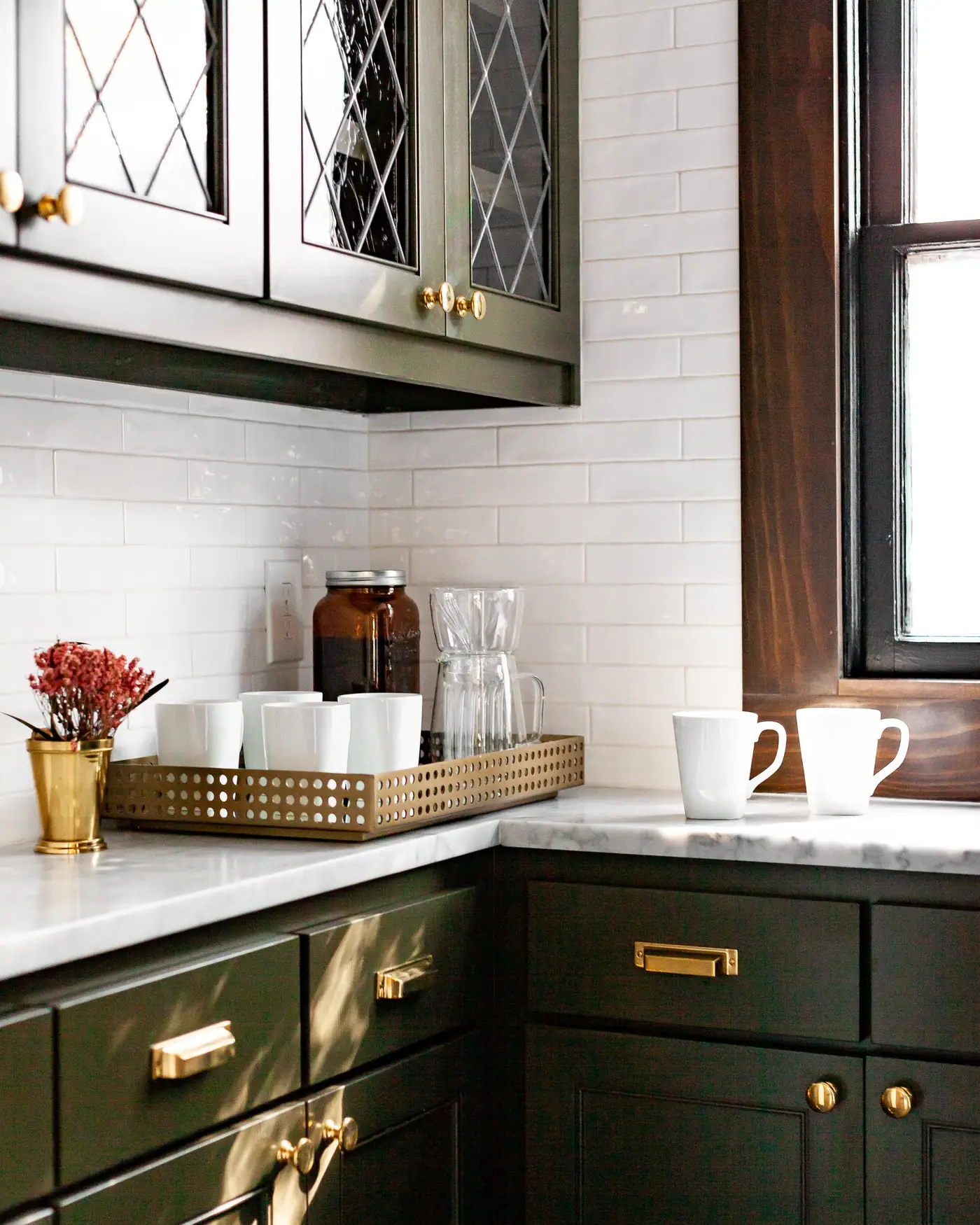 A coffee station in a remodeled kitchen with deep green cabinets, brass hardware and a white subway tile backsplash