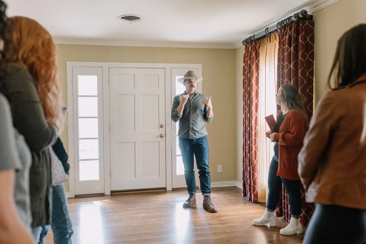 A builder in a hat talking a group of real estate agents through the empty front room of a house.