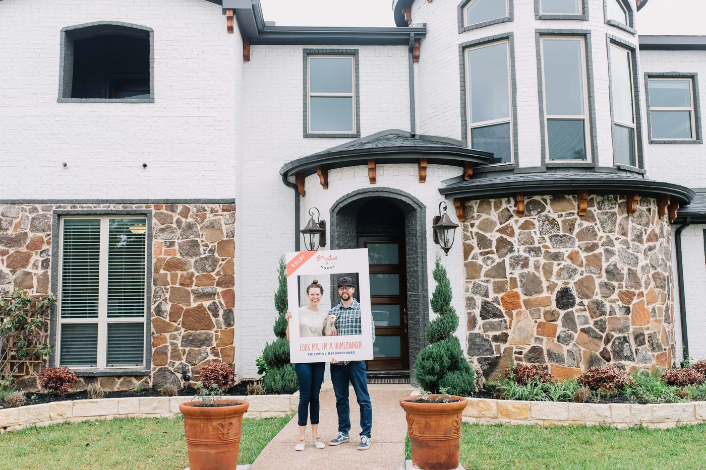 A couple holding a 6th Ave Homes “sold” frame outside the stone-arched entry of the home they just bought.