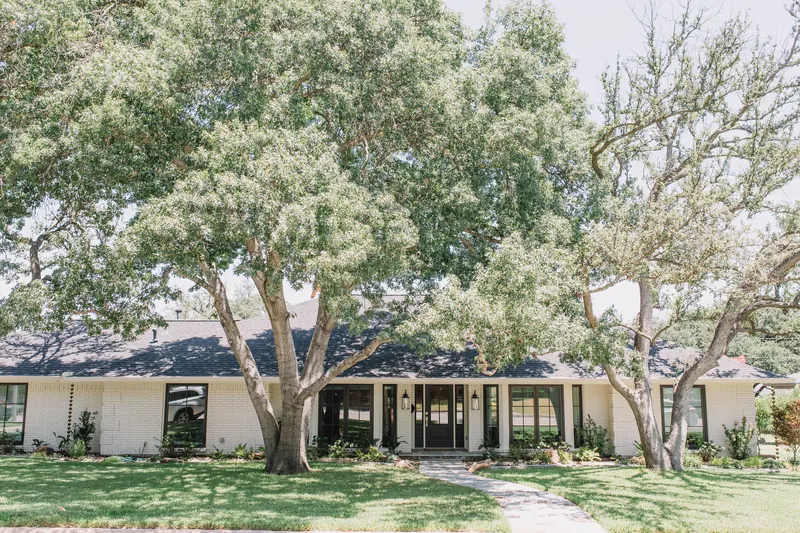 A white-brick Fort Worth ranch house under two live oaks, with black steel-look windows and a flagstone walk