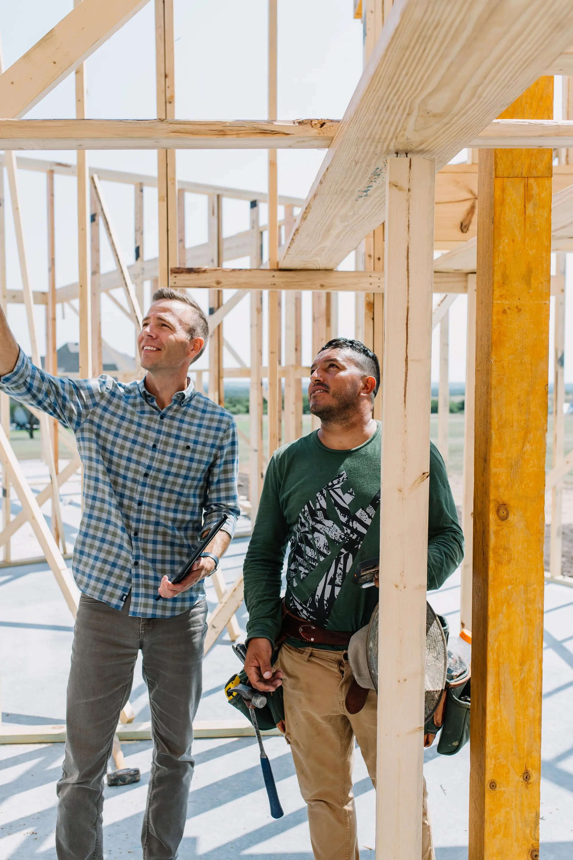 Two 6th Ave Homes builders inside the wood framing of a Fort Worth build, checking a beam against the plans
