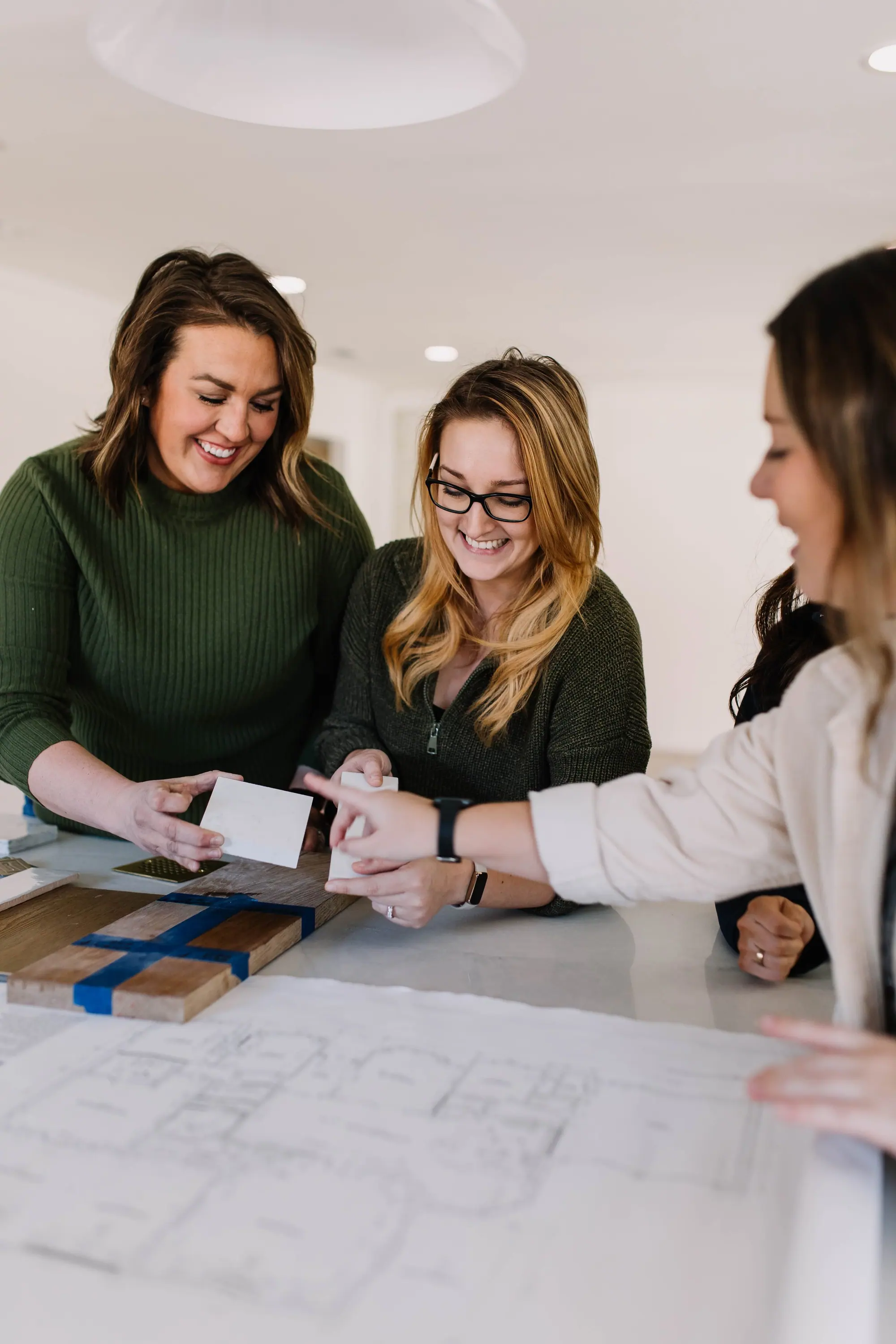 Three designers laughing over an unrolled floor plan and a board of wood and stone samples.