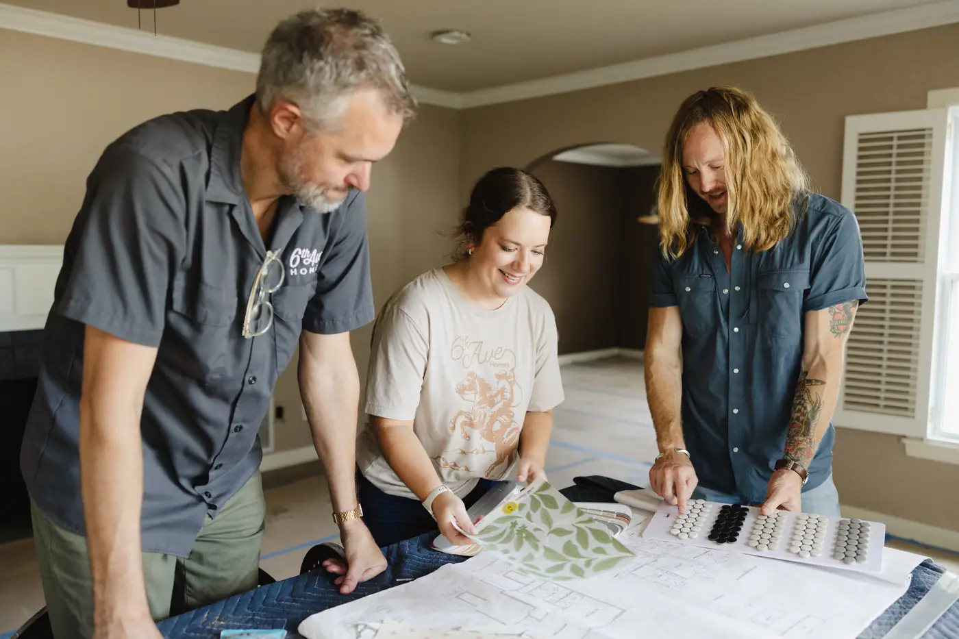 Three 6th Ave Homes team members laughing over a floor plan with tile and wallpaper samples