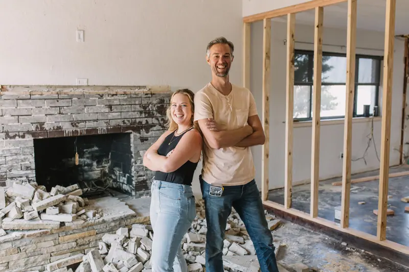 Homeowners grinning back-to-back in their mid-demo Fort Worth addition, exposed studs behind them