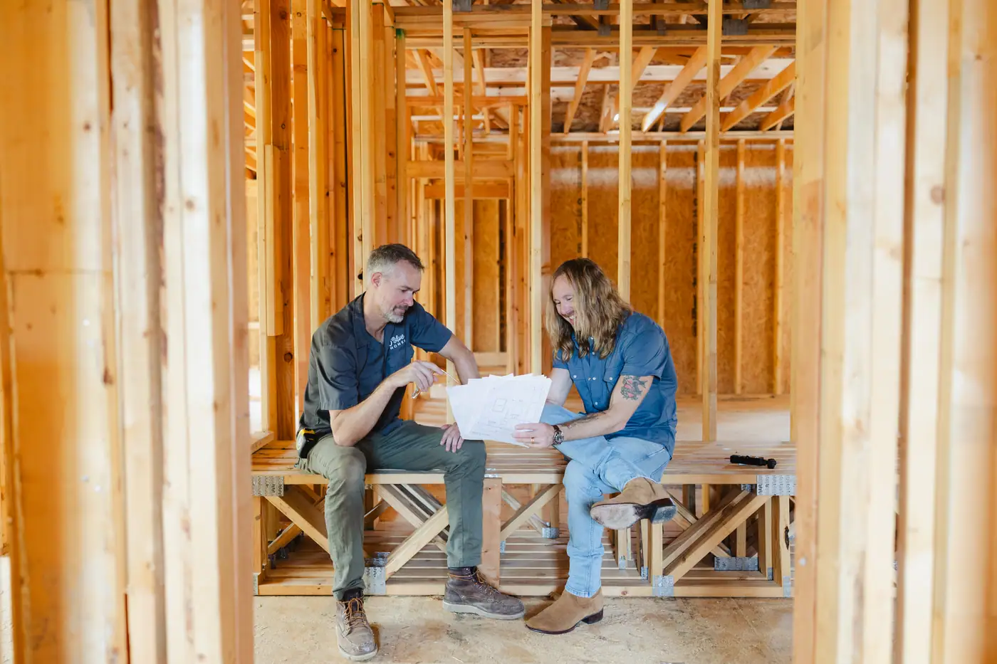 Two 6th Ave Homes team members reading a plan set on a bench inside the framing of a Fort Worth build