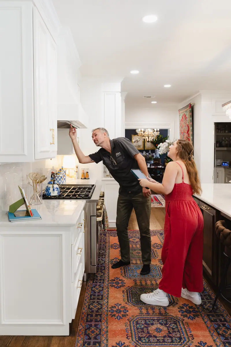 Two 6th Ave Homes team members on a walkthrough of a finished white kitchen: one reaches up to check the range hood while the other watches, holding an iPad