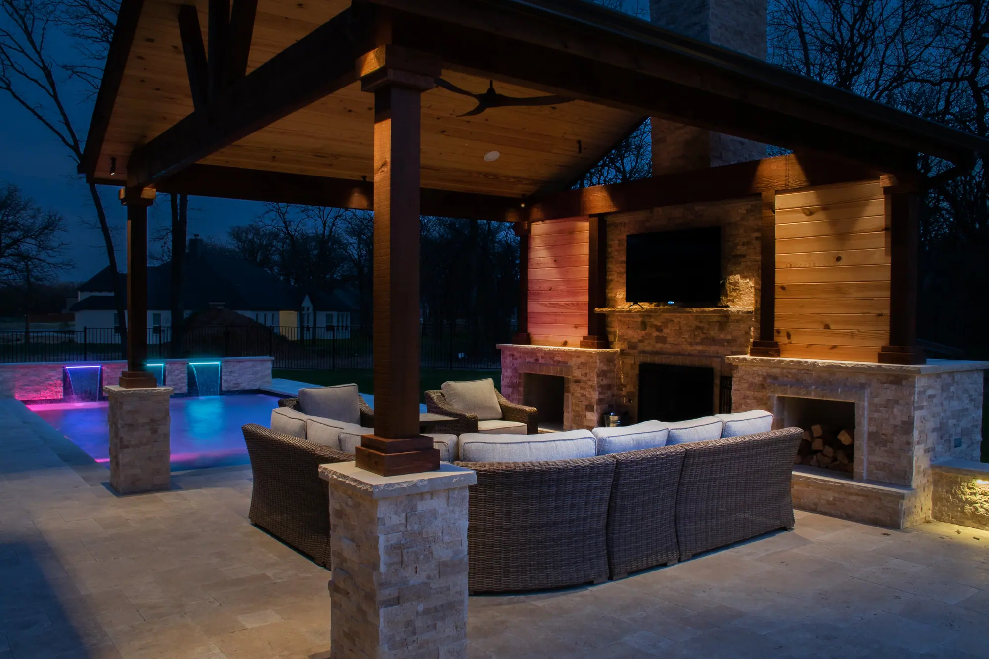 A gabled cedar pavilion at dusk with a stacked-stone fireplace, a curved sectional and a lit pool beside it