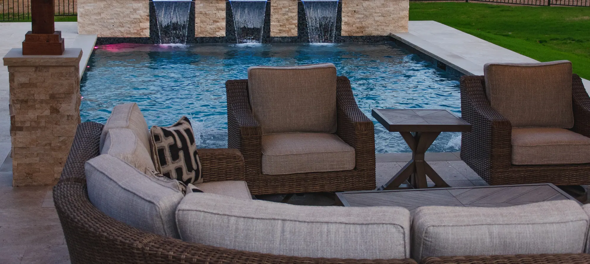 The view from under a freestanding pavilion: a stone column and cedar post, wicker chairs and a curved sectional facing a pool with sheer-descent waterfalls