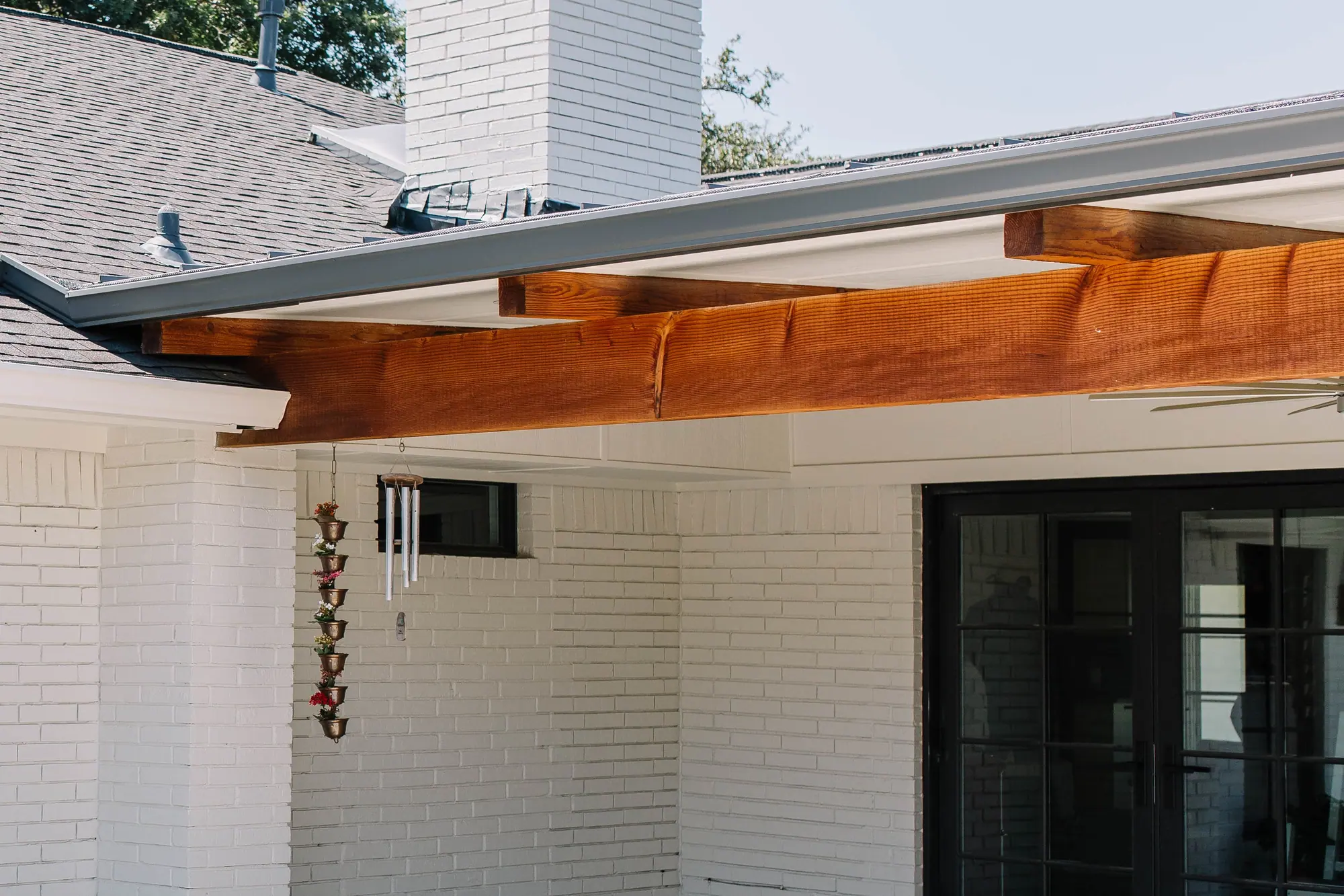 Where a new patio cover meets the house: its gutter running into the existing roofline, with cedar beams under the eave above a white-brick wall and French doors