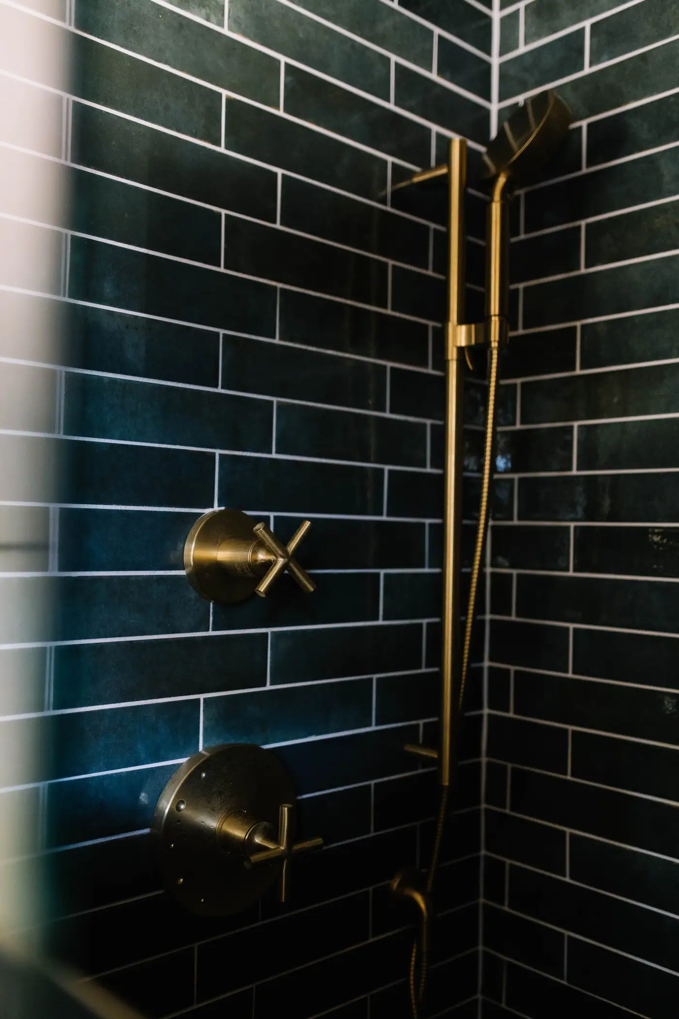 A shower wall of deep teal glazed subway tile with unlacquered brass cross-handle fixtures