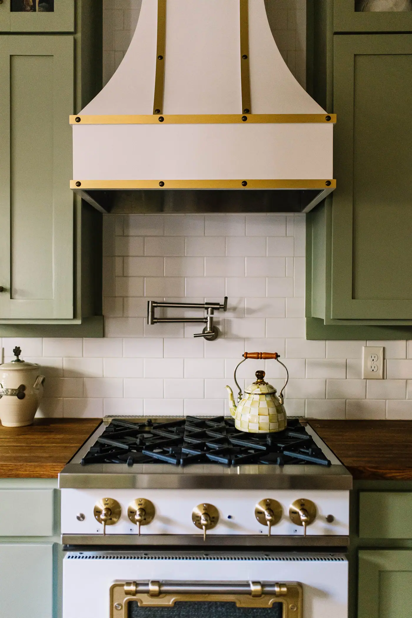 A kitchen with sage green cabinetry, a brass-banded plaster range hood, white subway tile and walnut counters