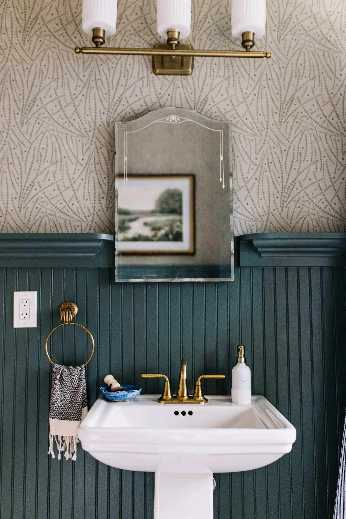 A powder room with deep teal beadboard wainscot, sprig-patterned wallpaper, an aged brass light and an antique bevelled mirror