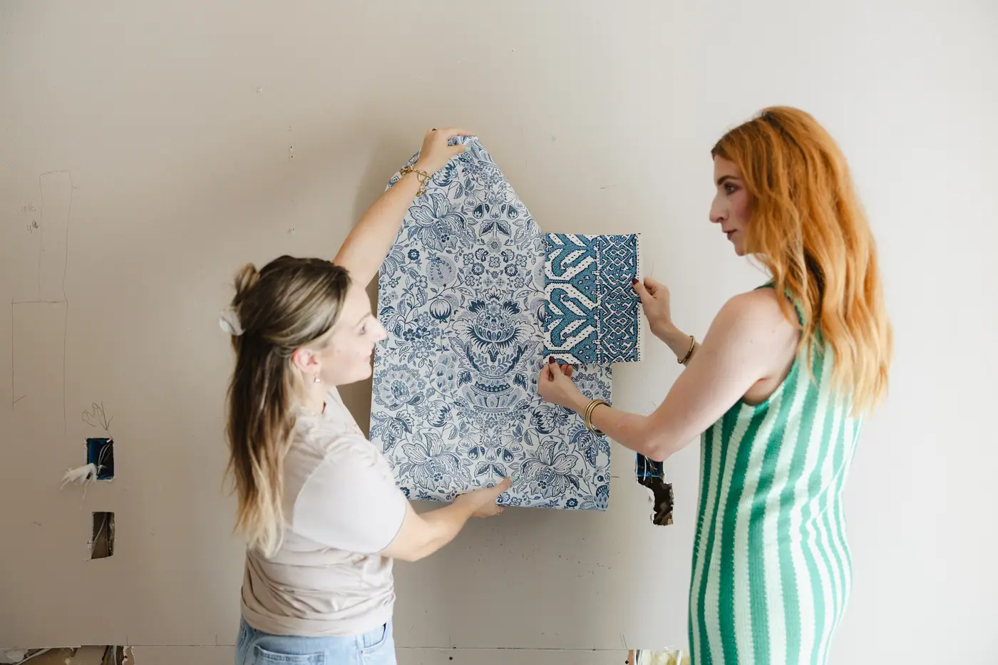 Two 6th Ave Homes designers holding blue wallpaper samples up to the wall of a Fort Worth home mid-renovation