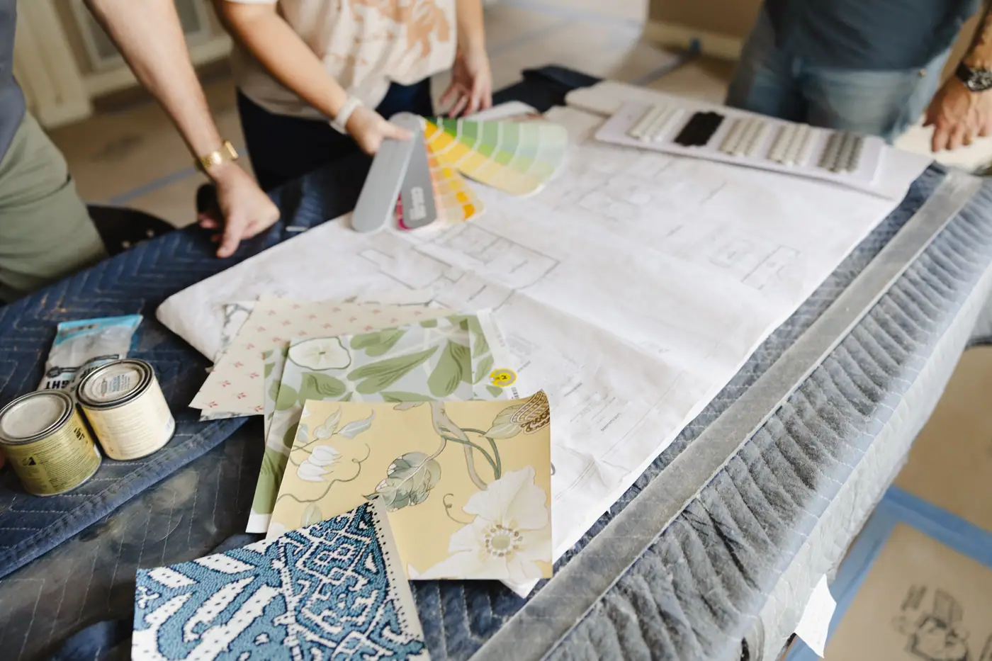 A floor plan on a padded table under a paint fan deck, wallpaper samples, two sample paint cans and a tile board, with hands at the edges.