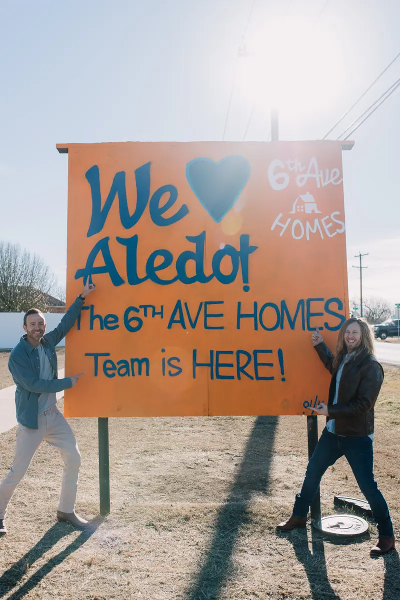 Two members of the 6th Ave Homes team pointing at a hand-painted sign reading “We love Aledo! The 6th Ave Homes Team is HERE!” on a dry winter lot