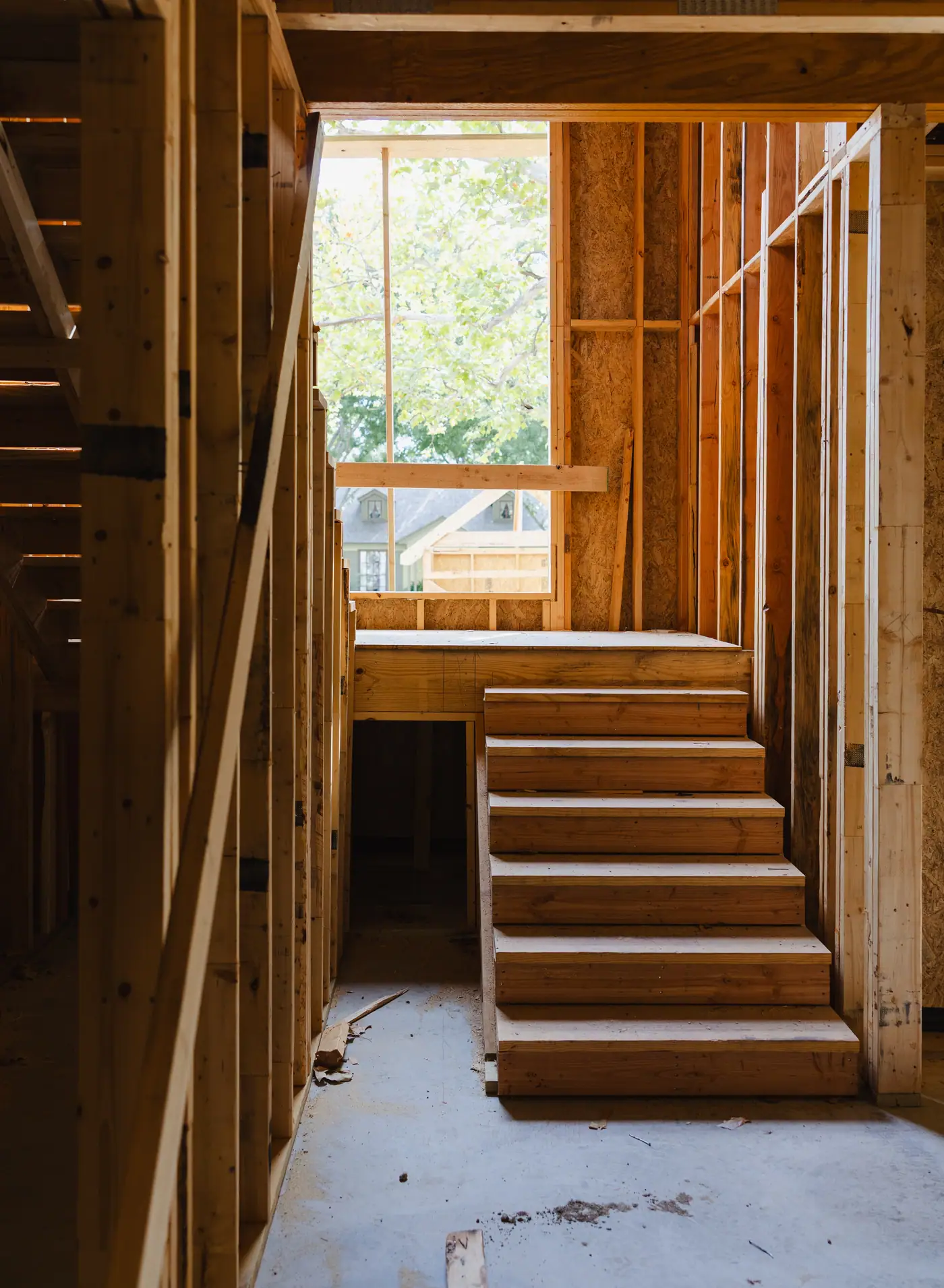 A raw framed staircase climbing to a landing under a tall window opening, inside a custom home under construction in Fort Worth
