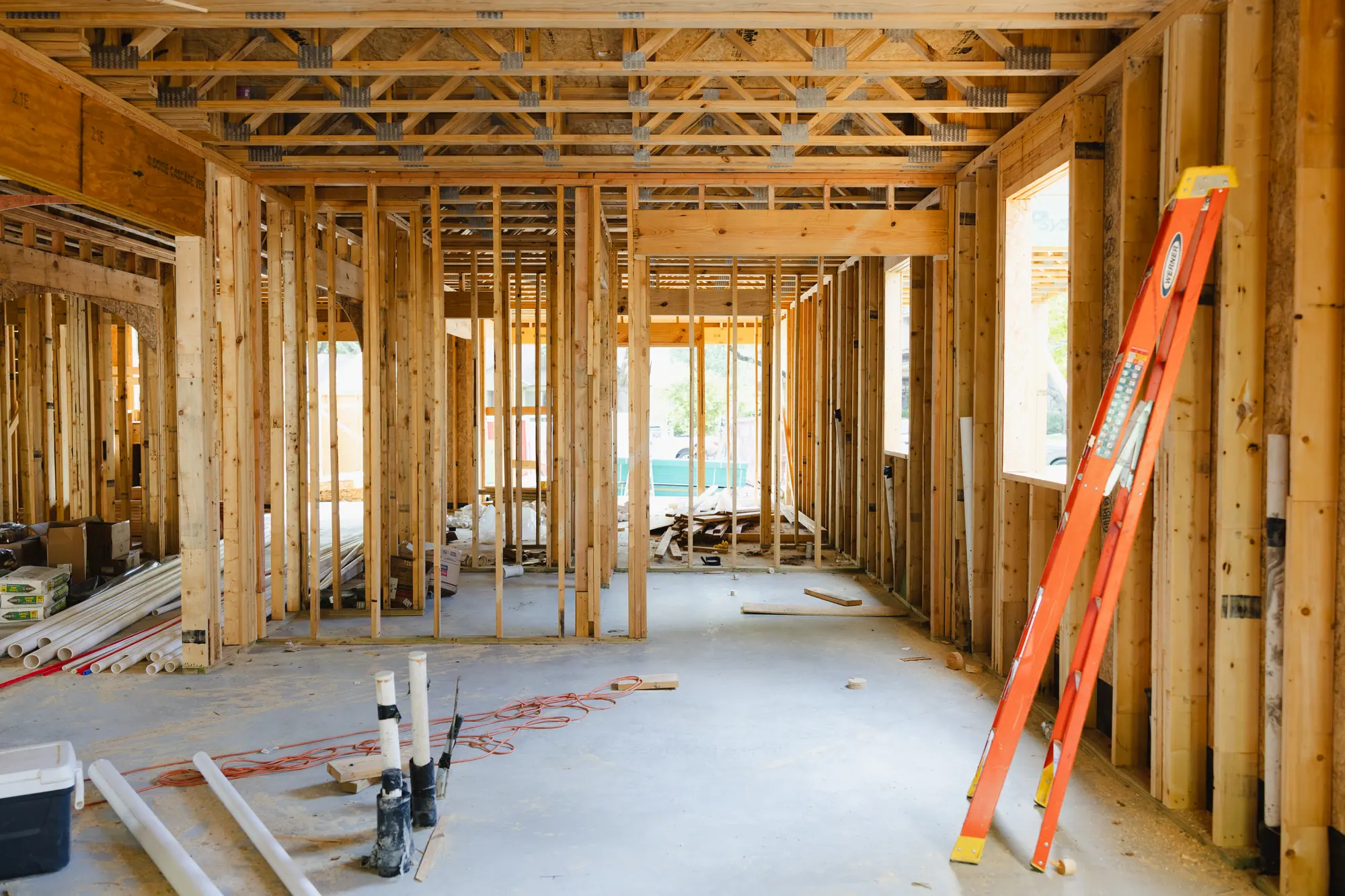 Inside a new house in framing: open stud walls, floor trusses overhead, plumbing pipes standing up from the slab, and a ladder against the wall.