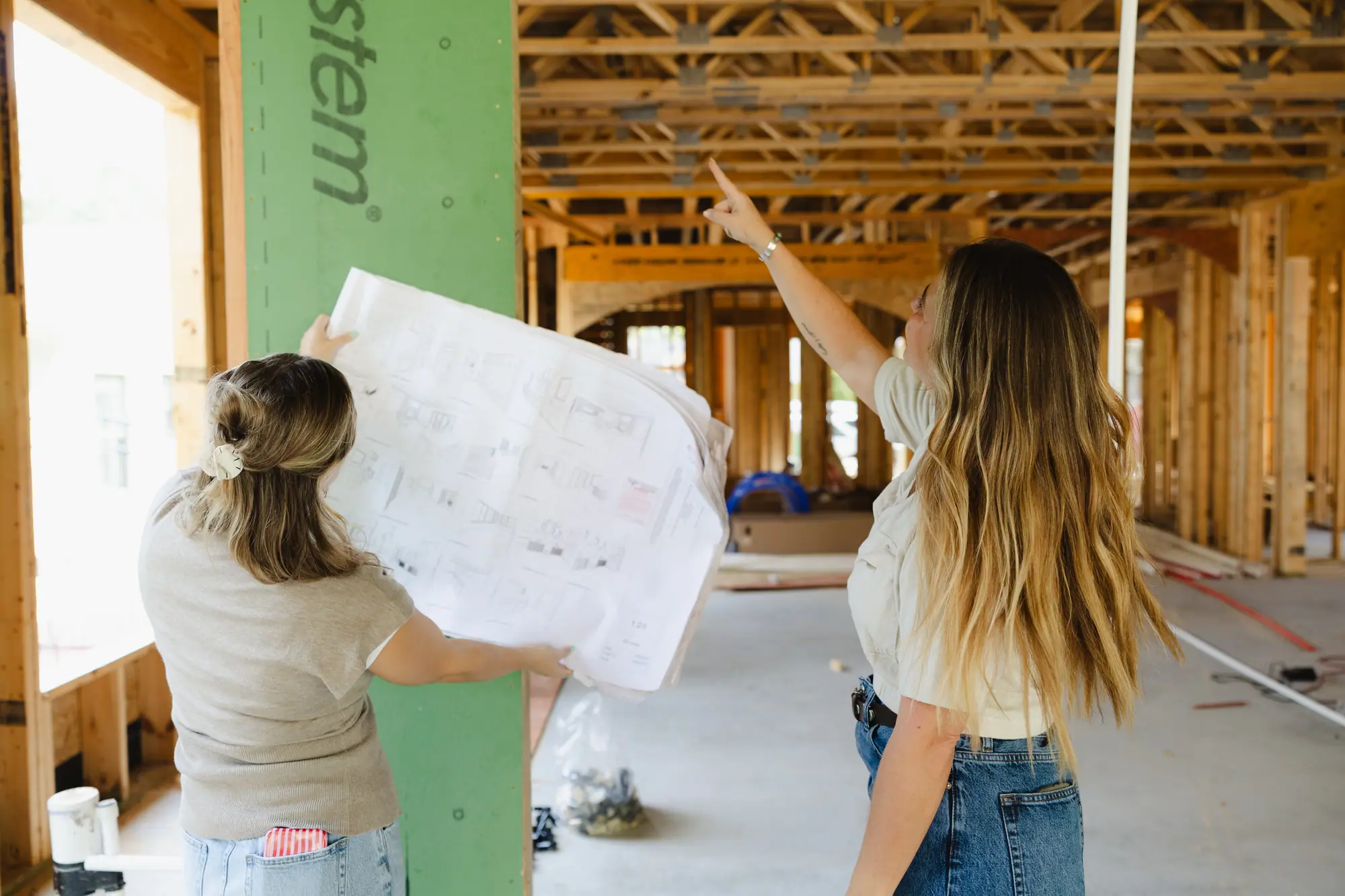 Two 6th Ave Homes team members hold up a sheet of a plan set inside a house in framing, one pointing up at the floor trusses