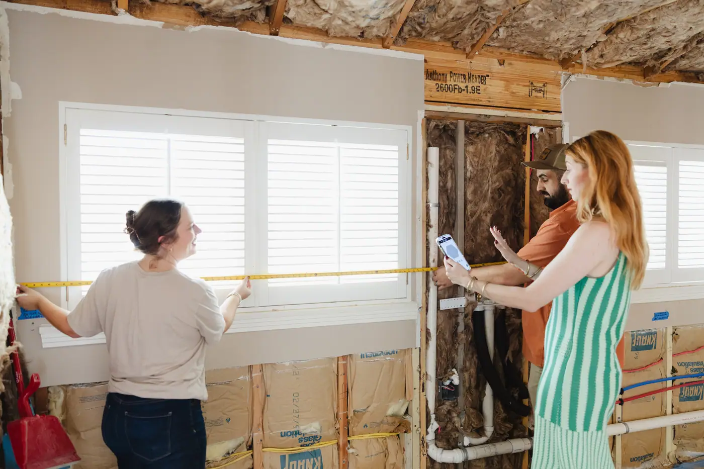 Three team members measuring inside a room opened to the studs, with the plumbing, wiring and insulation showing in the open wall.