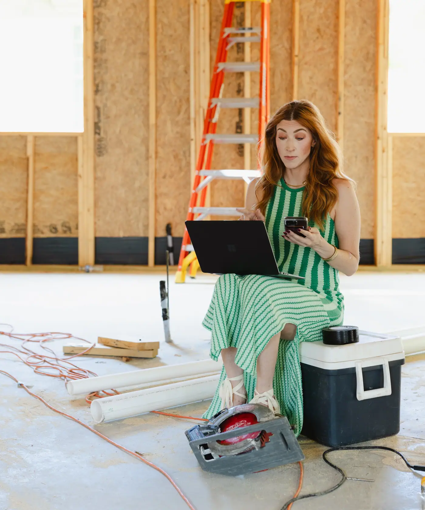 A 6th Ave Homes team member working on a laptop and a phone, seated on a cooler inside the framing of a custom home under construction