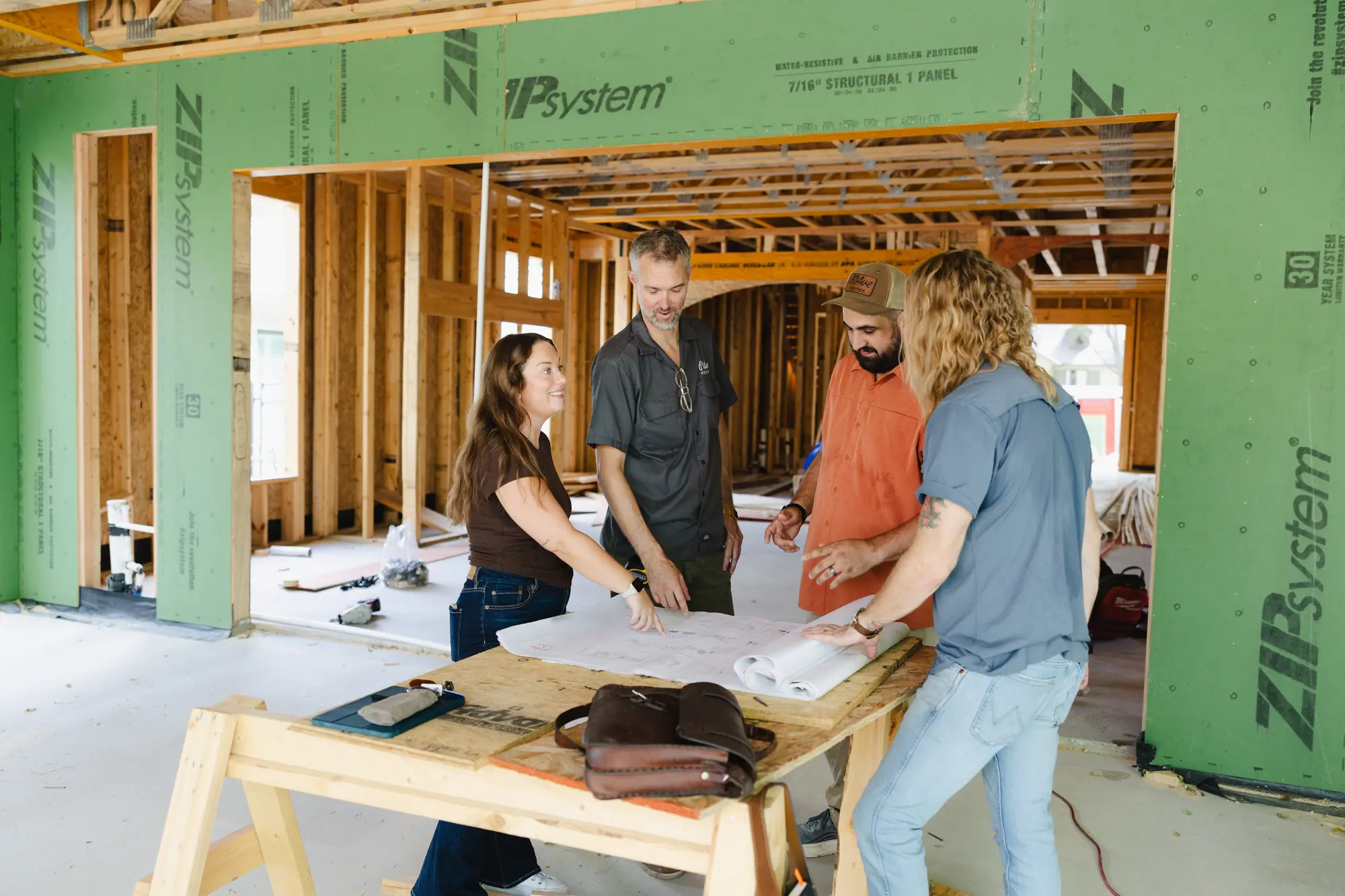 Four 6th Ave Homes team members talk over a plan set on a sawhorse table inside a house in framing