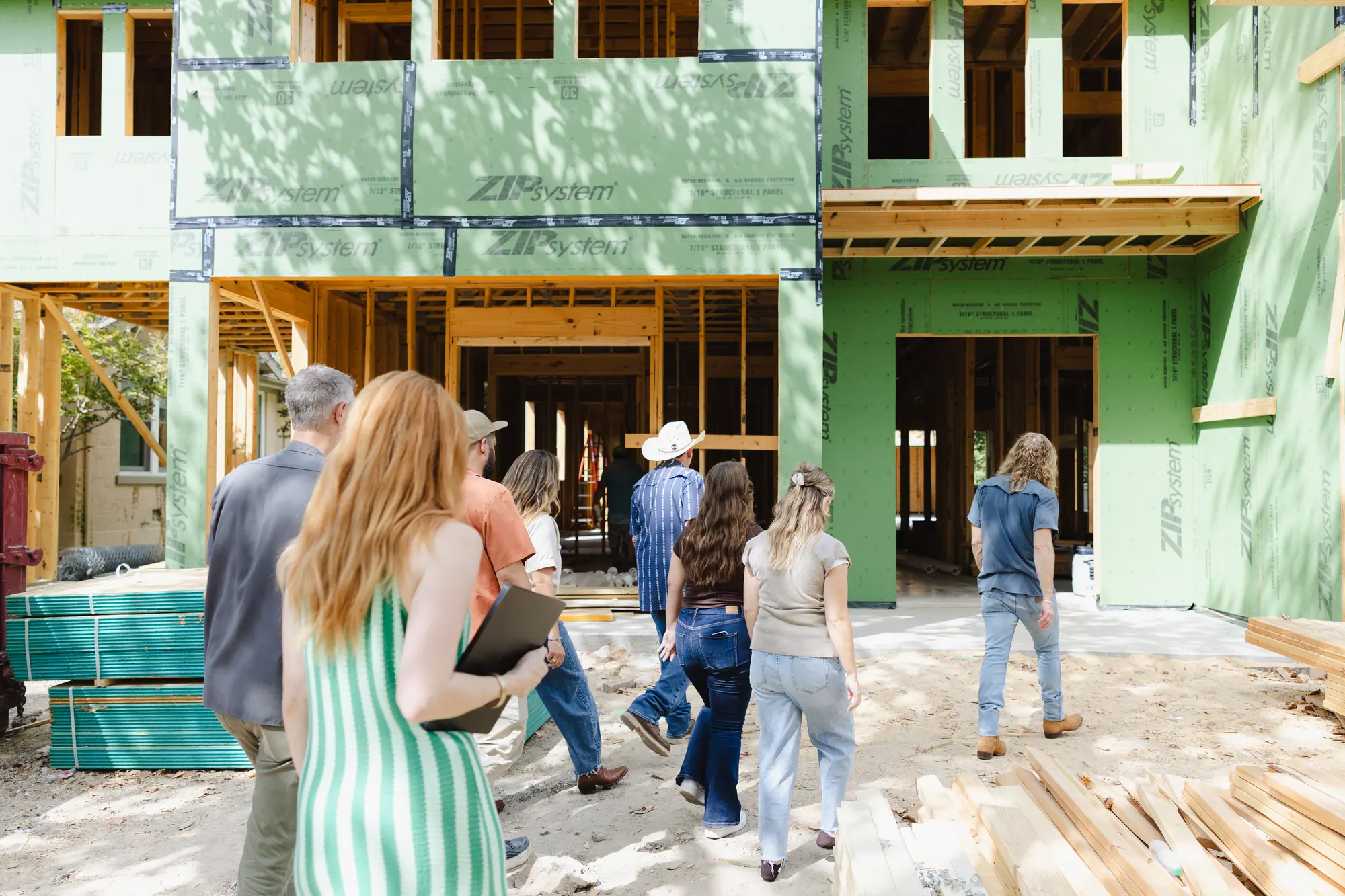 Our team, seen from behind, walking across a dirt lot toward a two-story new house in green sheathing with its framing still open.