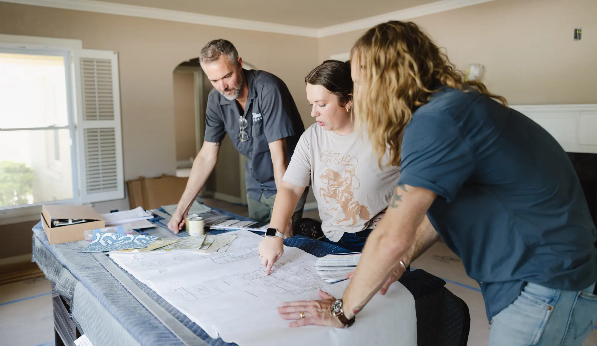 Three of our team going over a floor plan on a blanket-covered table, with wallpaper samples, sample paint cans and a paint fan deck beside it