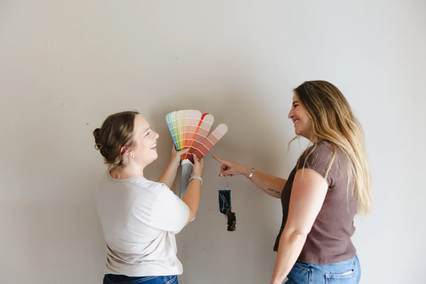 Two of our team laugh as one holds a fan of paint samples up to a bare wall and the other points to a coral shade.