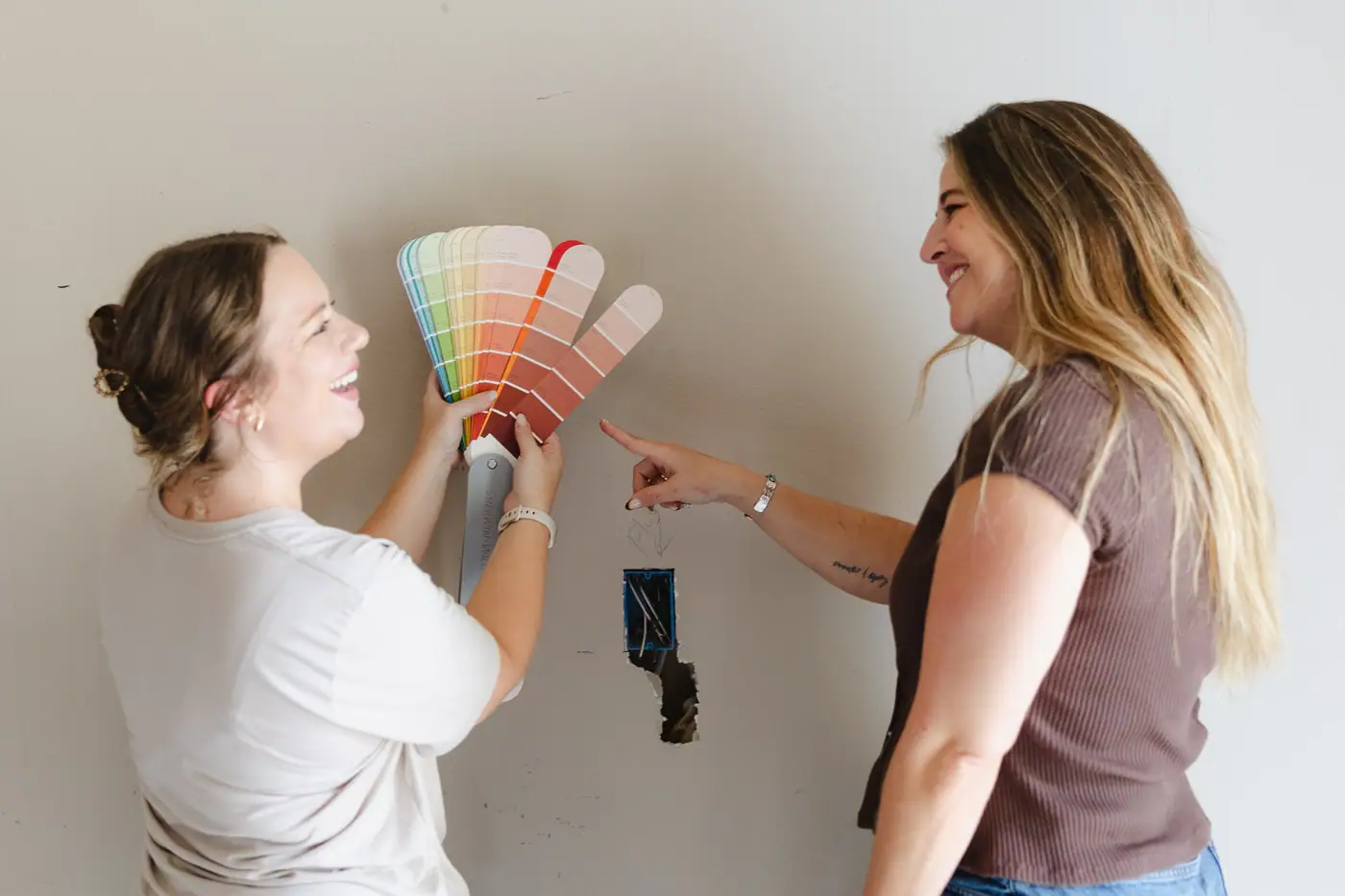 Two of our team laughing as one holds a fanned paint deck up to an unfinished wall and the other points to a coral shade