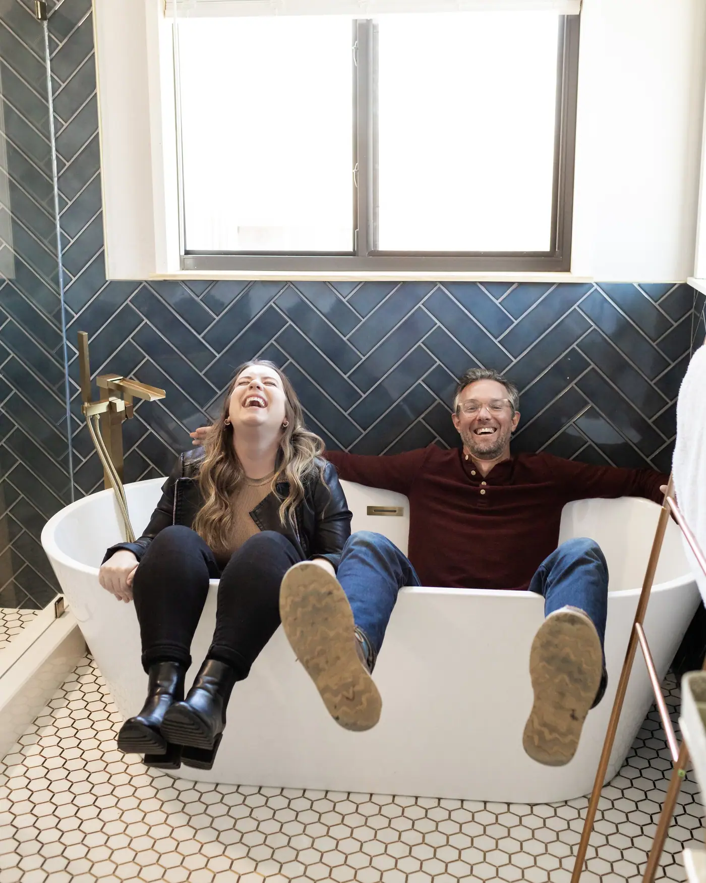 Two 6th Ave Homes team members sitting in a newly installed freestanding tub, laughing, against a navy herringbone tile wall