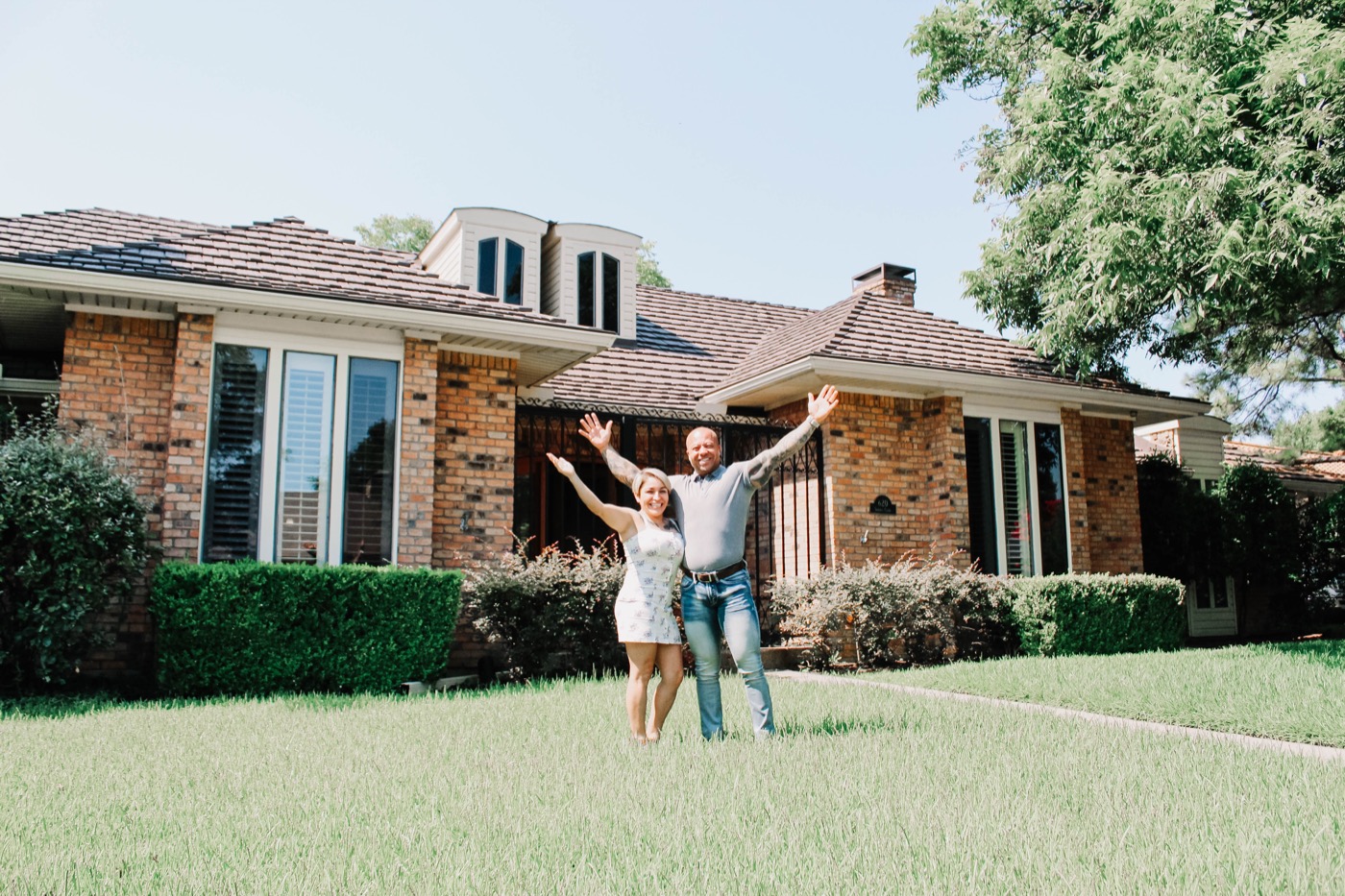 A Fort Worth couple throwing their arms up in front of the brick ranch they just bought, in full summer sun