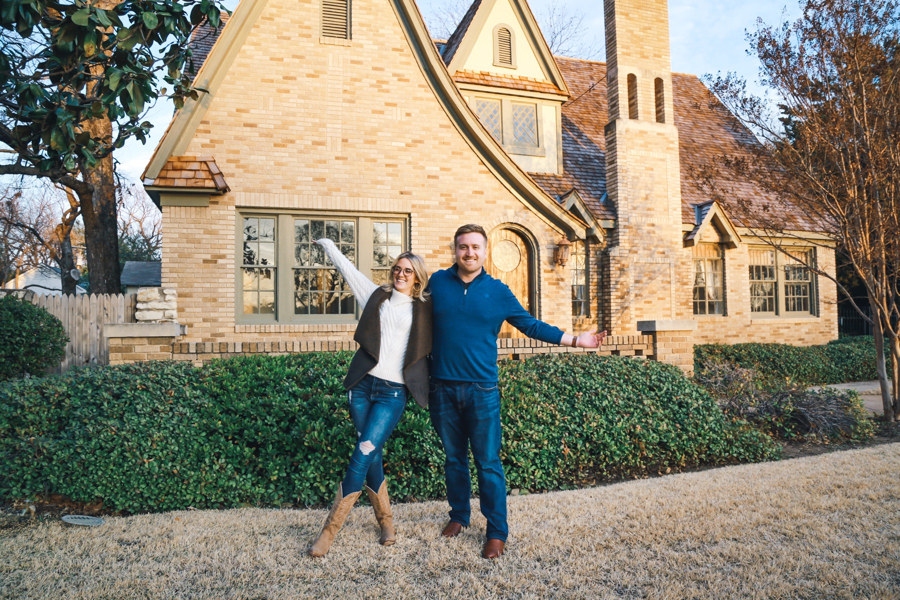 A Fort Worth couple, arms out, presenting the storybook brick Tudor they bought with 6th Ave Homes at golden hour