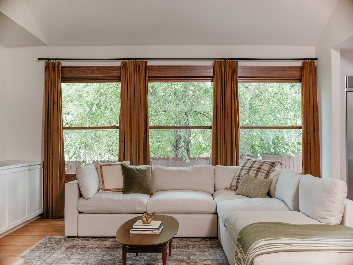 A great room opened to the kitchen in a Fort Worth whole-home remodel, with tall windows, rust velvet drapes, and a deep sectional