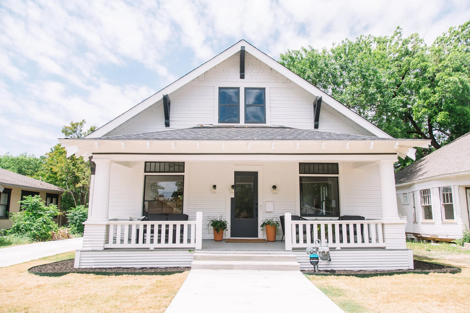 A freshly refreshed white Craftsman bungalow with a deep columned porch, black door, and mature landscaping in Fort Worth by 6th Ave Homes