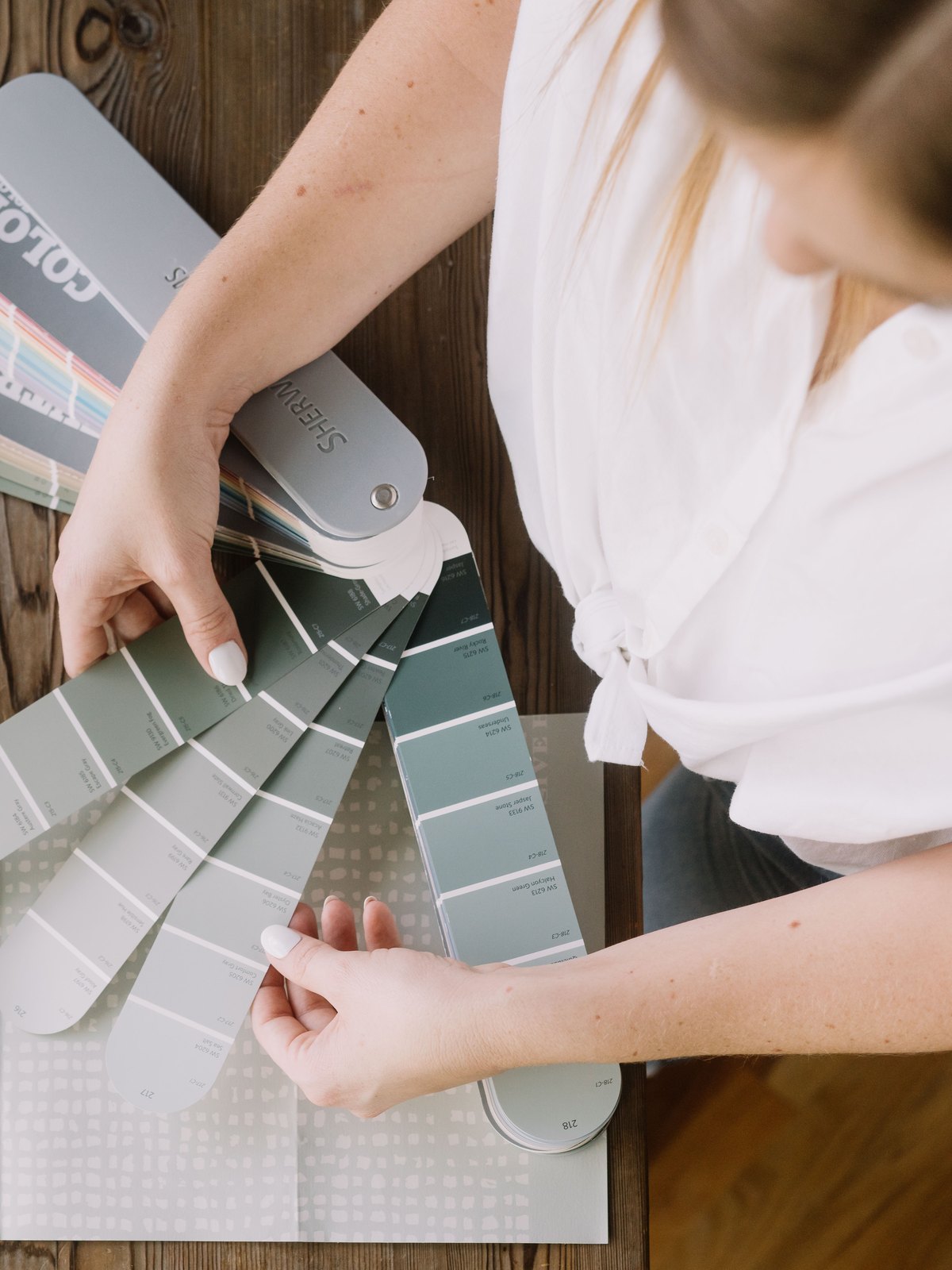 An interior designer opening a fan of sage and grey paint chips across a walnut table, with tile and wood samples laid out around it