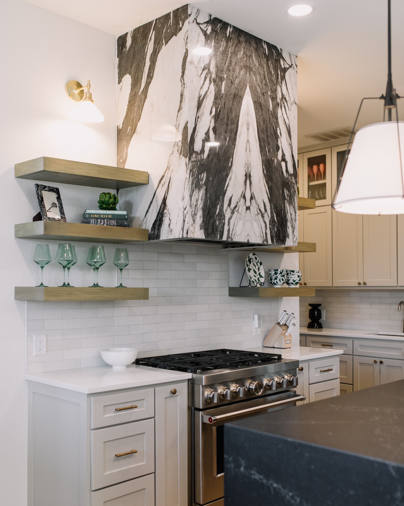 A dramatic book-matched marble waterfall range hood over a pro gas range, oak shelves, and a black stone island in a Fort Worth kitchen