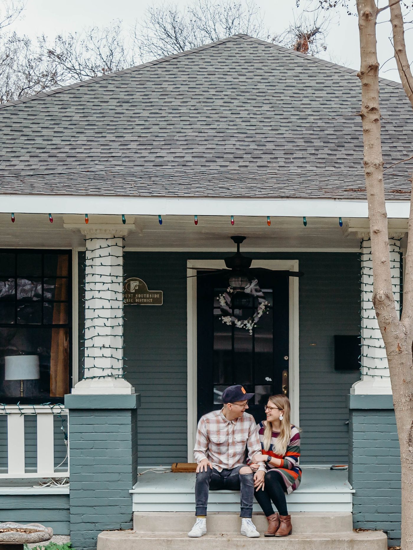 A couple sitting on the front steps of their historic Fairmount bungalow in Fort Worth under holiday lights