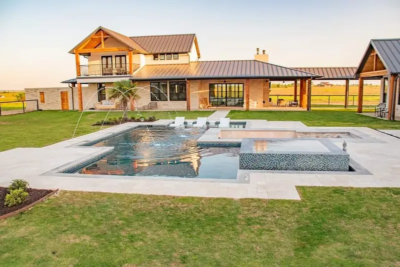 A geometric pool and pebble spa with deck jets at golden hour behind a modern farmhouse