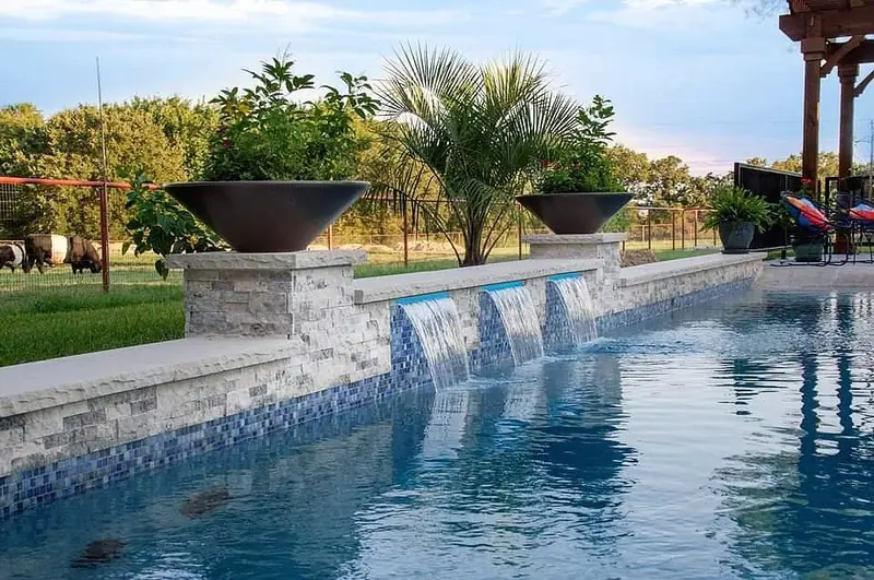 A stacked-stone wall with sheer-descent waterfalls and fire bowls above a Fort Worth pool