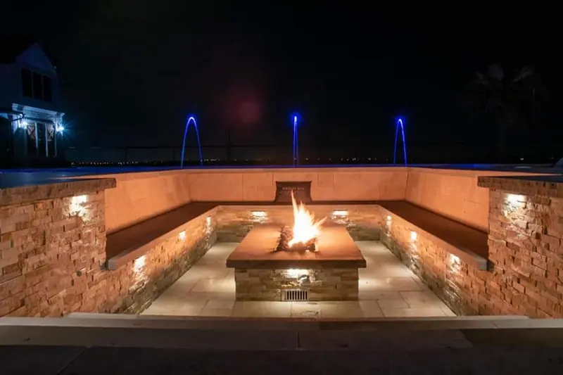 A sunken stone fire lounge at night with a burning fire table and lit water features behind