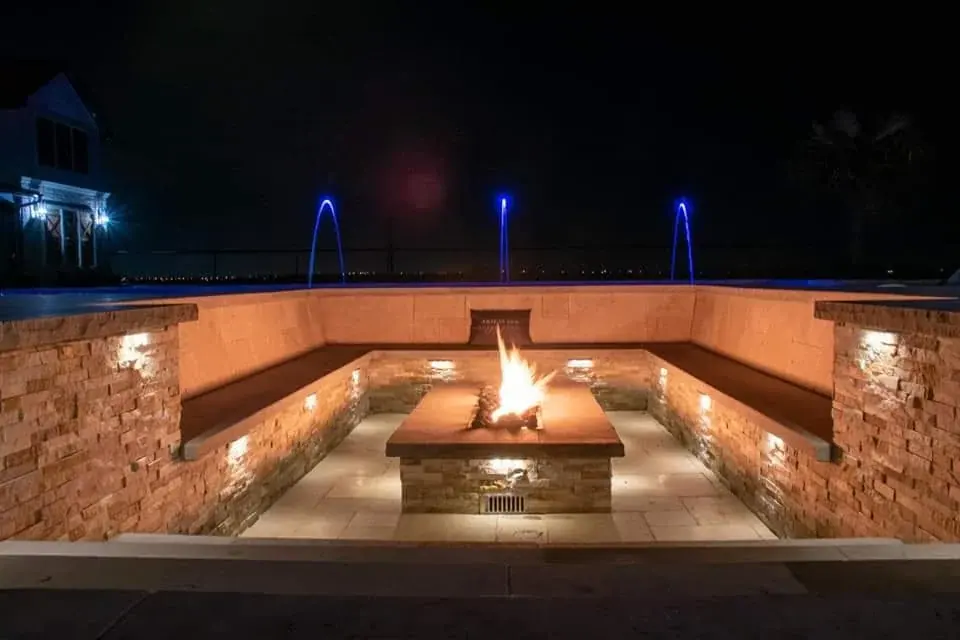 A sunken stone fire lounge at night with a burning fire table and lit water features behind