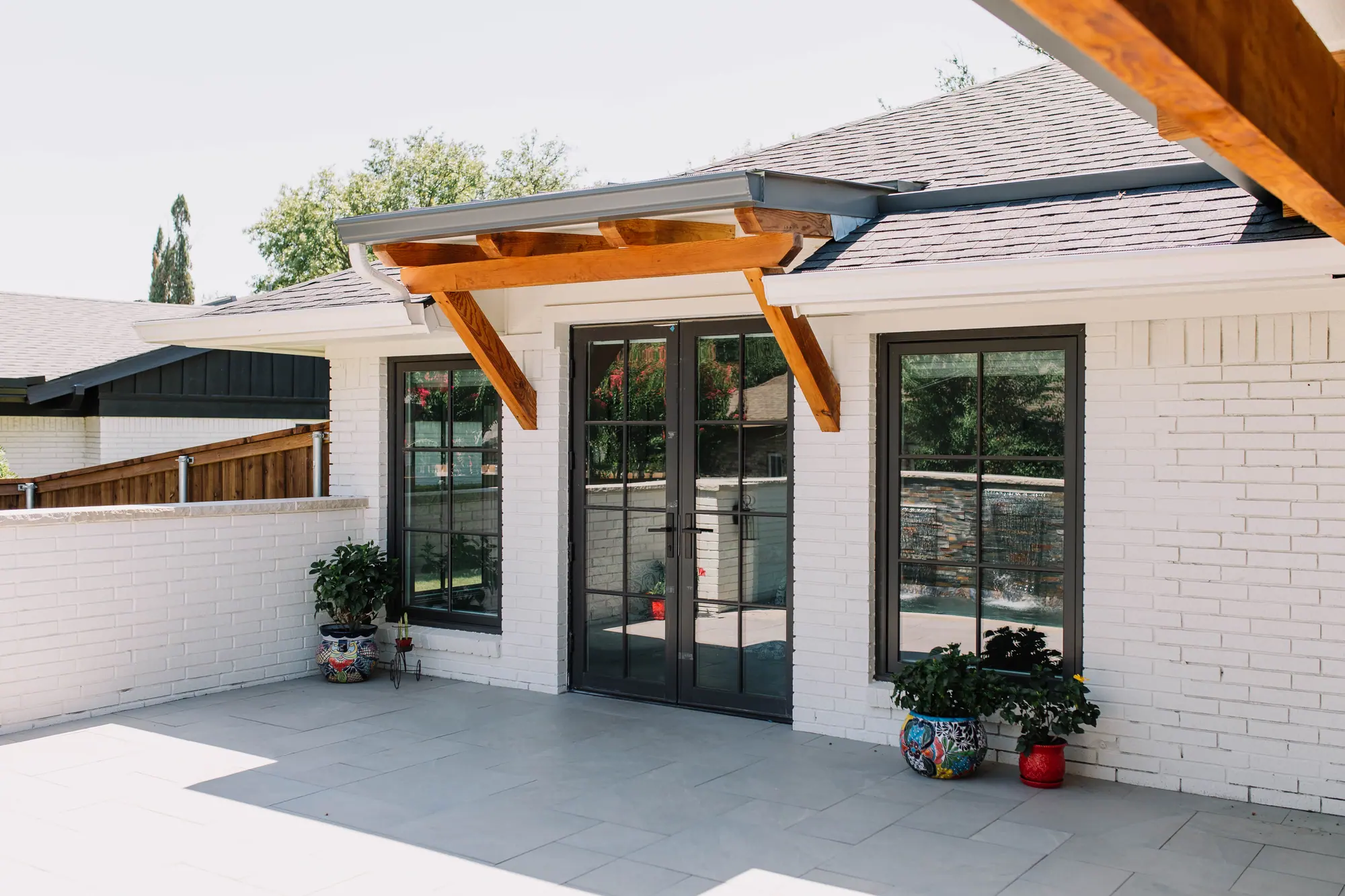 The back of the Wedgwood house seen from under the outdoor kitchen: French doors under a cedar-bracketed awning, black-framed windows and painted white brick