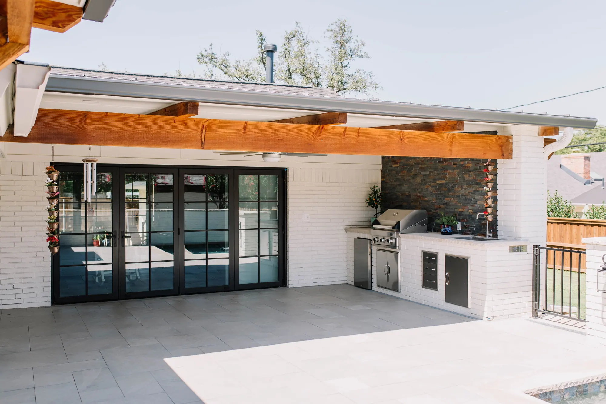 A covered patio in Wedgwood: black-framed glass doors into the house on the left, the outdoor kitchen on the right under the same cedar-beamed roof