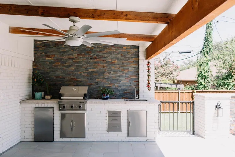 A white-brick outdoor kitchen with built-in grill and beverage fridge under a cedar-beamed patio cover in Fort Worth