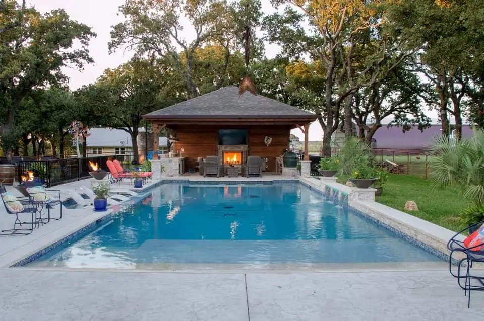 A long rectangular pool with in-water loungers, a hip-roofed cedar cabana with a lit fireplace at the far end under oaks