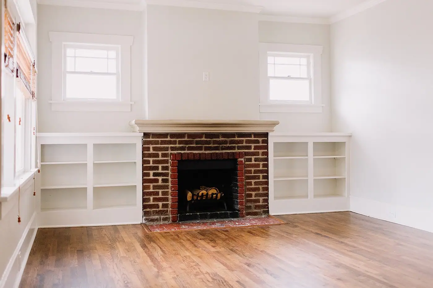 An empty room with a bare oak floor, a red-brick fireplace and empty white built-ins — a house between owners