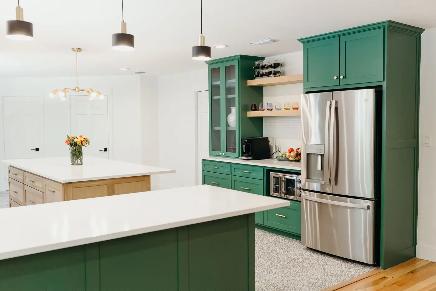 Open kitchen with emerald cabinetry and a white oak island, three pendants over a quartz peninsula and flowers on the counter.