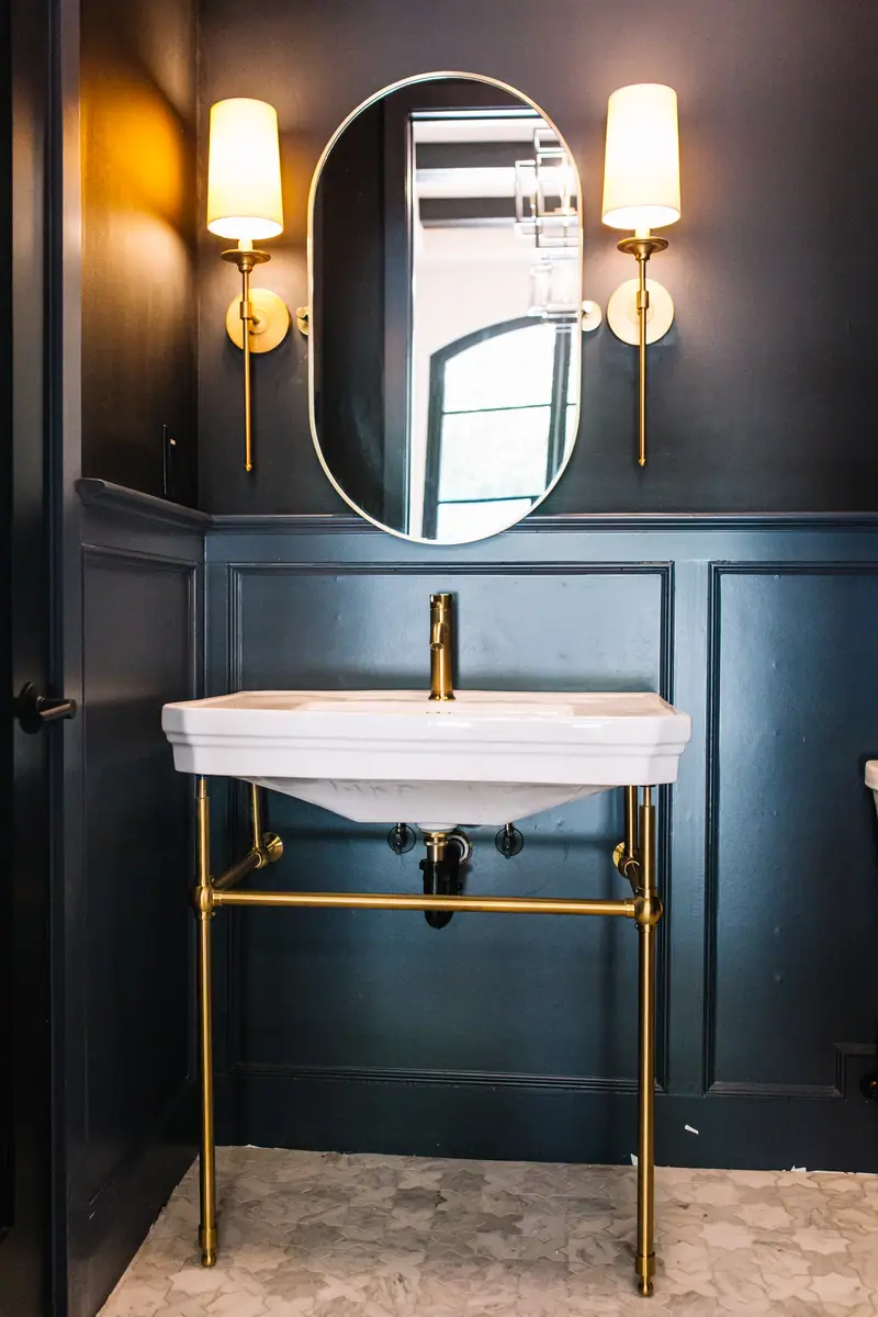 A dark, panelled bath with a brass console sink, an oval mirror and brass sconces